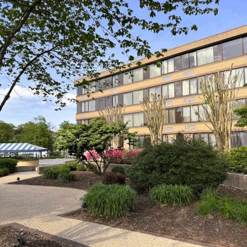 A building with multiple windows surrounded by trees and shrubs, with a pathway leading toward a structure with a blue-striped canopy.