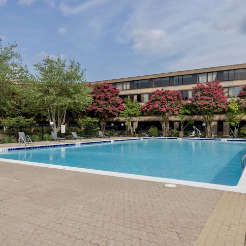 An outdoor swimming pool with crystal-clear water, surrounded by trees and flowers, in front of a multi-story building under a partly cloudy sky.