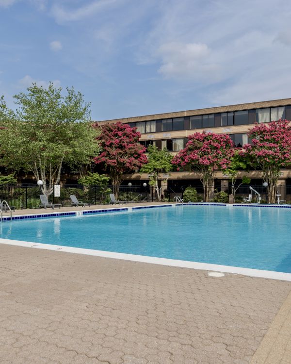 An outdoor swimming pool with crystal-clear water, surrounded by trees and flowers, in front of a multi-story building under a partly cloudy sky.