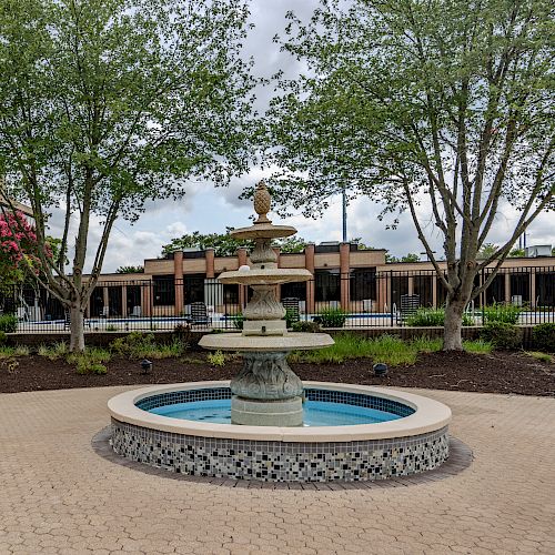 An outdoor courtyard features a central tiered fountain, surrounded by trees, plants, and a brick pathway. A building is visible in the background.