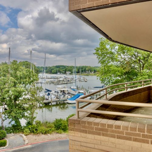 The image shows a marina with boats docked, a balcony, and greenery under a cloudy sky.
