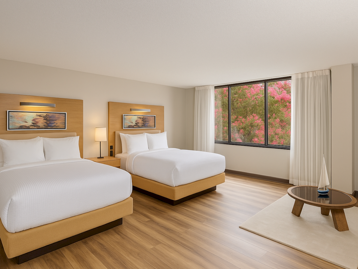 The image shows a modern hotel room with two beds, a large window with curtains, hardwood floors, and a small table with a decorative piece.