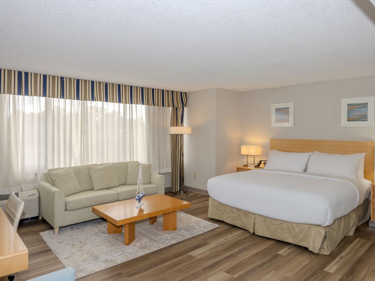 A bright, modern hotel room features a large bed, a sofa, a coffee table, side lamps, a desk, and large windows with a striped valance.