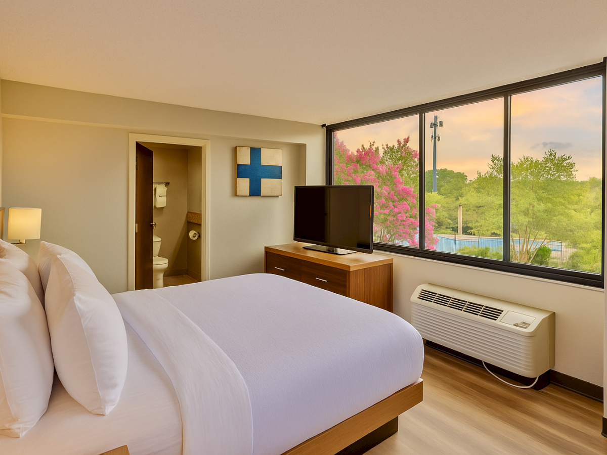 A hotel room with a neatly made bed, TV, large window, air conditioner, and a bathroom. There's a scenic view outside the window.
