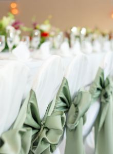 A long banquet table with white-covered chairs, each adorned with green bows, set with glassware, napkins, and flower centerpieces, ready for an event.