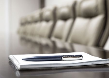 A pen rests on a notepad placed on a polished conference table, with empty chairs in the background, suggesting a meeting setting.