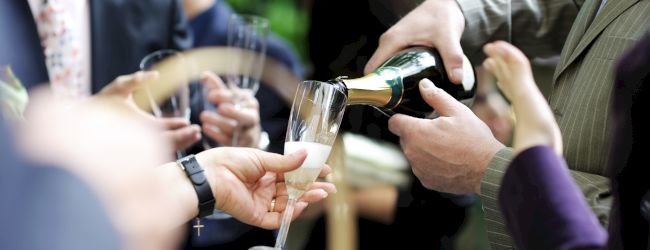 A group of people dressed in formal attire is gathered, with a person pouring champagne into a glass at what appears to be a social event.