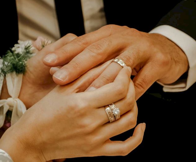 Two hands with wedding rings are seen, one hand placing a ring on another. They might be part of a wedding ceremony or a similar celebration.