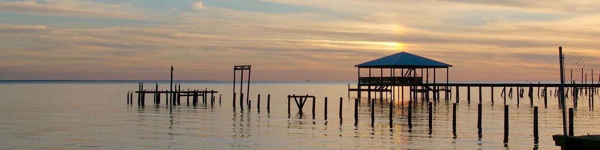 A serene waterscape at sunset with a wooden pier, a small dock, and a boathouse on stilts, reflecting on calm water.