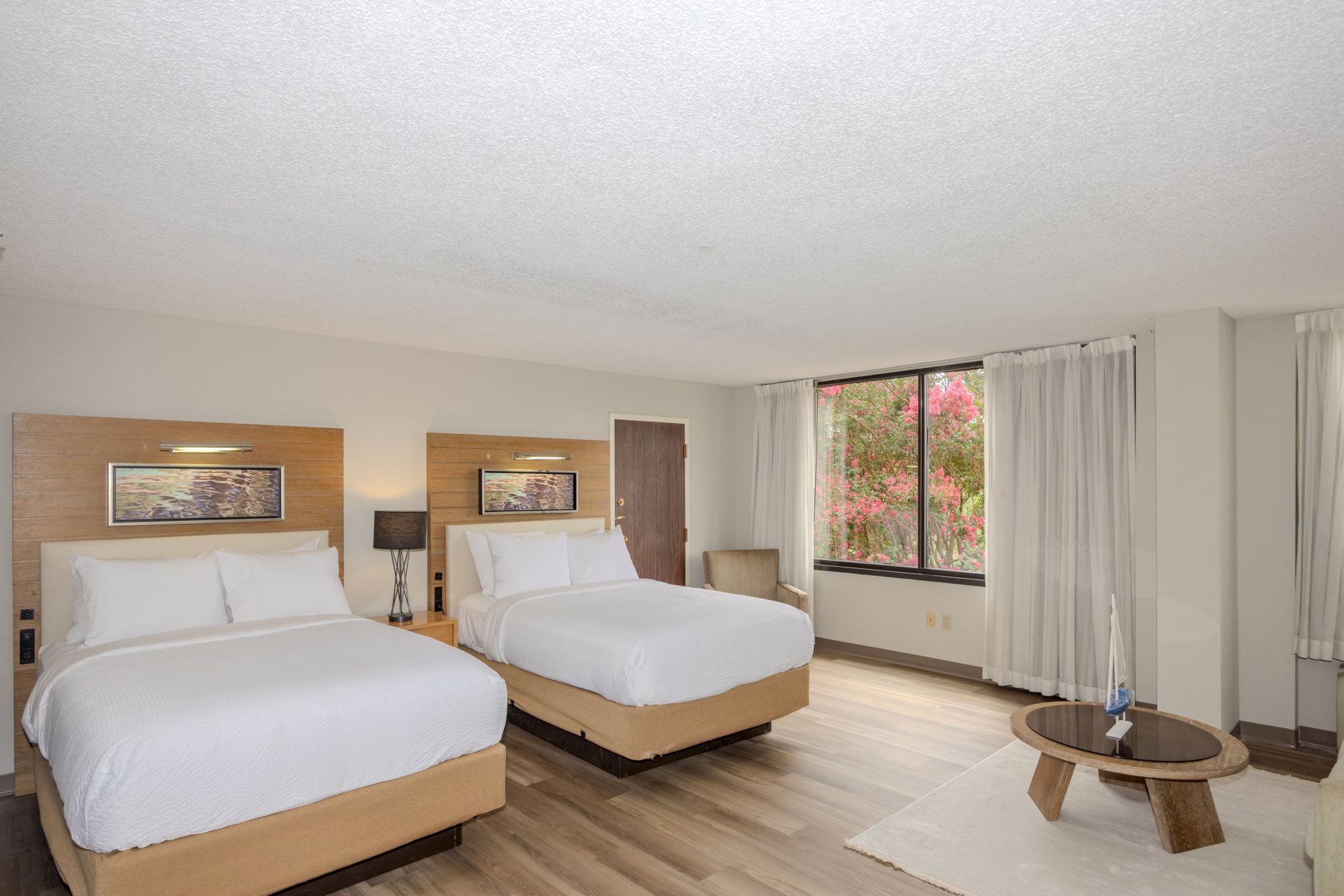 A modern hotel room with two double beds, a lamp, a large window, an armchair, and a small round table.