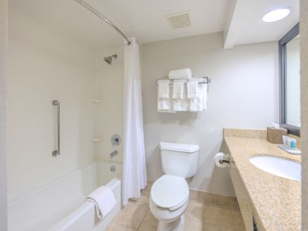 The image depicts a clean, modern bathroom with a bathtub and shower, a toilet, a sink with a large countertop, and towels on a rack.