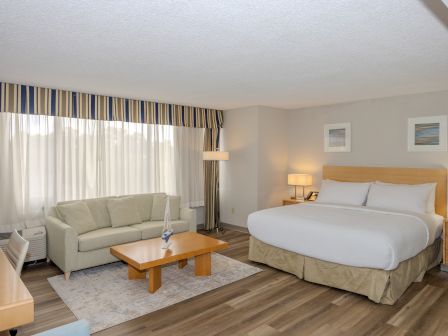 A hotel room with a bed, a sofa, a desk, two lamps, a coffee table, and framed pictures on the wall. The room features wooden flooring.