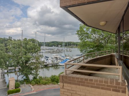 A dock with sailboats and a view from a brick building's balcony, surrounded by greenery and cloudy skies, creating a peaceful waterside scene.