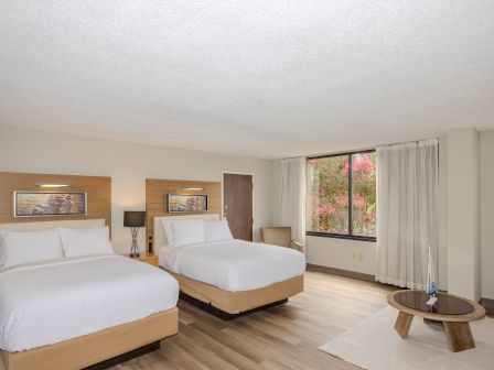 A modern hotel room with two double beds, a window with curtains, a side chair, a round table, and a painting on the wall above the beds.