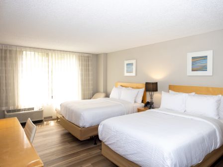 The image shows a well-lit hotel room with two double beds, a desk, a table lamp, abstract art on the wall, and large windows with curtains.