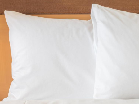 The image shows two white pillows placed side by side on a bed with a light-colored wooden headboard in the background.