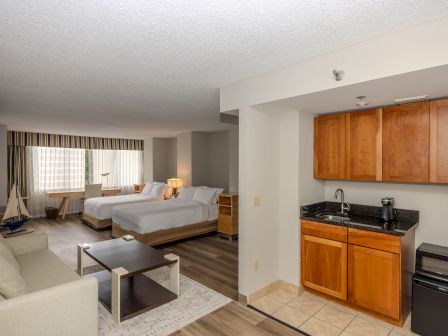 The image shows a hotel room with a kitchenette, two beds, a sofa, a coffee table, and nautical-themed decor with a large window in the background.