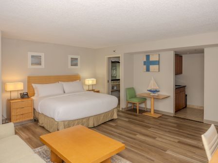 A modern hotel room with a large bed, bedside tables, lamps, a small dining area, a kitchenette, framed artwork, and wooden floors.