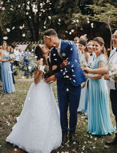 A bride and groom kiss outdoors, surrounded by bridesmaids in blue dresses and guests throwing flower petals in celebration.