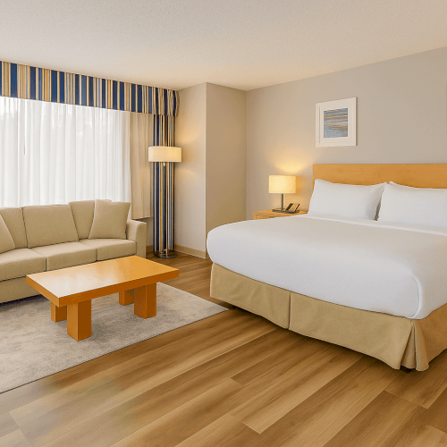 A modern hotel room with a large bed, sofa, coffee table, and lamps on wooden flooring, featuring a window with striped curtains.