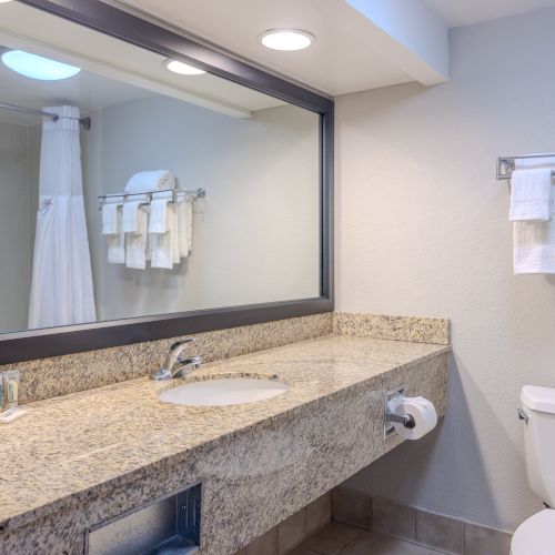 A clean and modern bathroom with a large mirror, granite countertop, sink, toilet, and neatly folded towels on a rack.