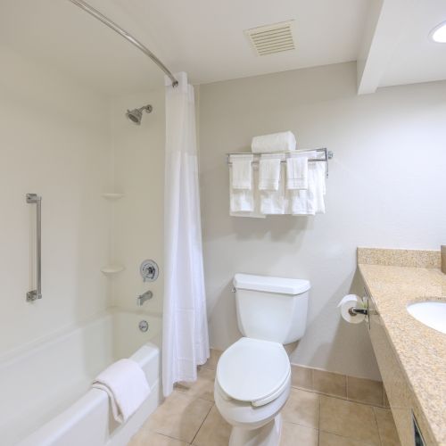 A neat bathroom with a bathtub, shower curtain, toilet, sink with granite countertop, mirror, towel rack with towels, and toiletries on the counter.