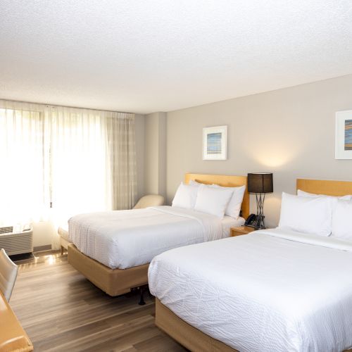 A well-lit hotel room with two neatly made double beds, a desk, chair, and bedside lamps, featuring a large window with curtains and framed art on the walls.