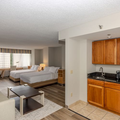 This image shows a hotel room with two beds, a kitchenette, a sofa, a coffee table, and a large window with curtains at the far end of the room.