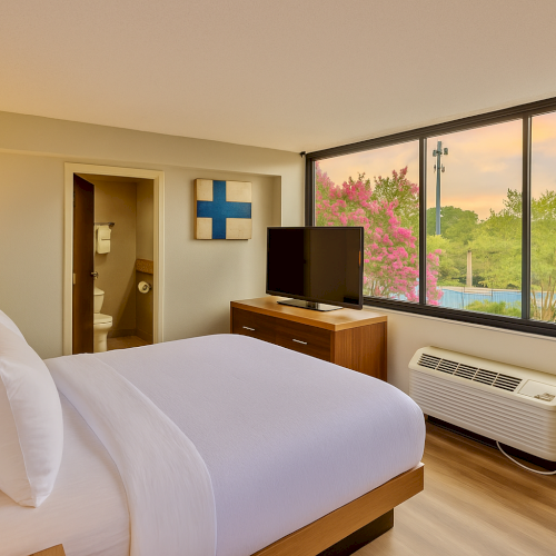 A modern hotel room with a bed, TV, and window view of trees and sky. A bathroom is visible through an open door.