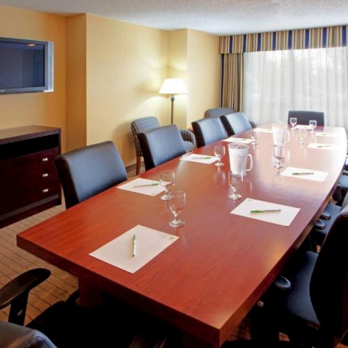 The image shows a modern conference room with a long table, chairs, a wall-mounted TV, notepads with pens, water glasses, and natural lighting.