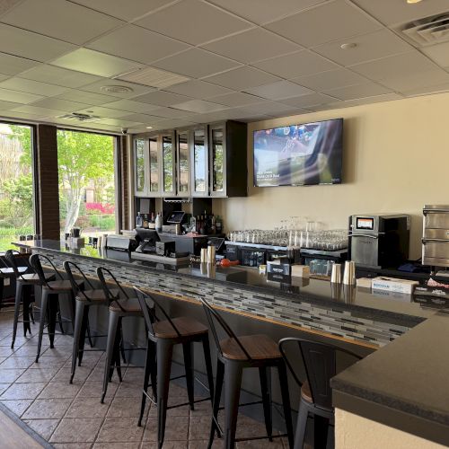 A modern bar with high chairs, a TV, glasses, a pizza oven, and scenic windows showing greenery outside.