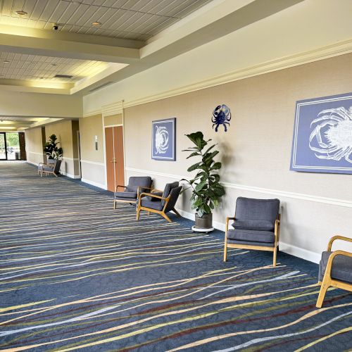 A carpeted hallway with chairs, plants, and crab-themed artwork on the wall, leads to glass doors with a view outside.