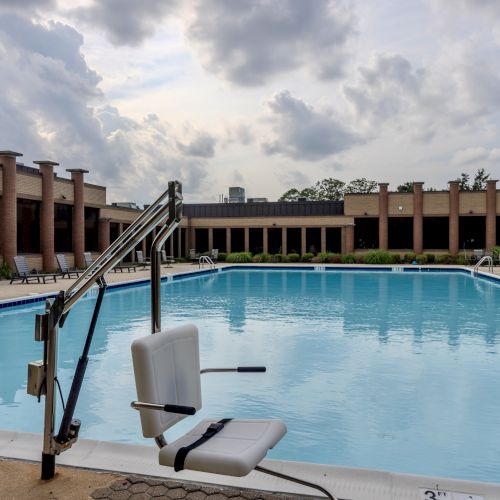 An outdoor swimming pool with lounge chairs and a mechanical lift seat next to it, surrounded by a brick building under a cloudy sky.