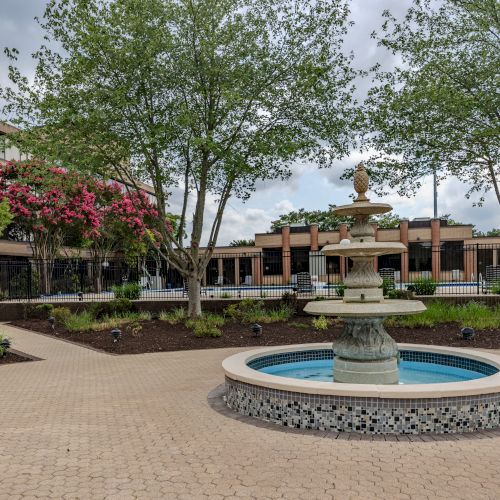 The image displays a courtyard with a central water fountain, surrounded by trees and lush landscaping near a multi-story building.