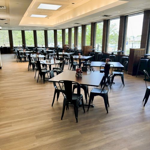 The image shows an empty restaurant with wooden floors, black tables, chairs, and large windows providing natural light.