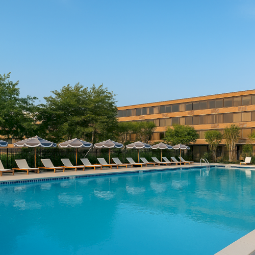 A large outdoor pool with lounge chairs and umbrellas, situated in front of a multi-story building and surrounded by trees.