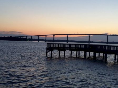 The image shows a sunset over a body of water with a bridge in the background and a pier in the foreground.