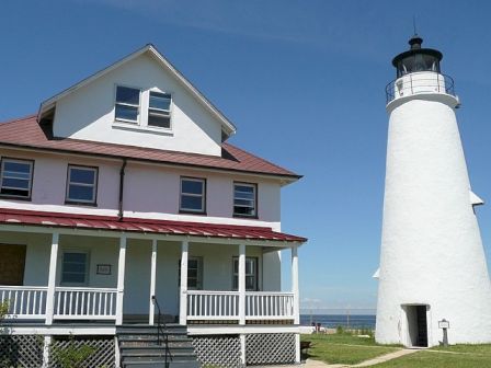 This image features a white house with a porch adjacent to a tall, white lighthouse under a clear blue sky on a grassy area near the coastline.