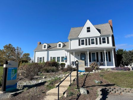 A white, two-story house with a front porch, garden, and sign near the entrance, under a clear blue sky. The house has dormer windows.