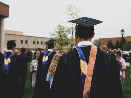 The image shows graduates in caps and gowns at an outdoor ceremony, surrounded by people and buildings.