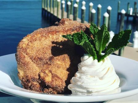 The image showcases a slice of dessert topped with whipped cream and mint leaves, set against a scenic background of water and piers in a marina.