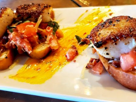 The image shows four seared scallops on toast, garnished with diced tomatoes and arugula, served on a white plate with a yellow sauce.