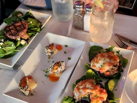 Four plates of food and two mason jars with drinks on a table. Grilled dishes and appetizers served on rectangular plates adorned with greens.