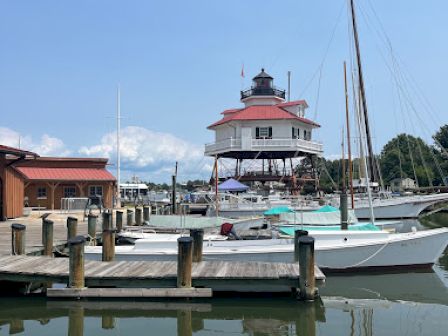 The image shows a marina with boats docked and a prominent lighthouse with a red roof in the background under a clear sky.