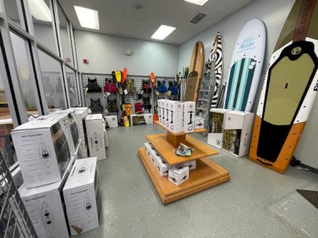 The image shows a store section with various paddleboards, water sports gear, life vests, and boxes of items displayed on shelves.