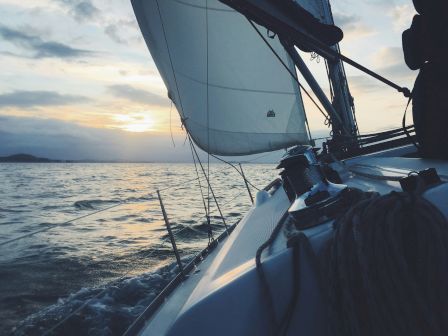 A sailboat gliding on the water at sunset, with visible sails, ropes, and a serene sea reflecting the evening sky.