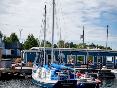 A small sailboat is docked at a marina, with a blue building and trees in the background under a partly cloudy sky.