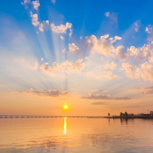 A serene sunrise over calm water, with scattered clouds illuminating the sky and a bridge in the distance, creating a peaceful ambiance.