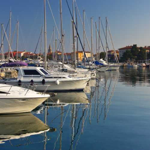 The image shows a marina with numerous sailboats and yachts docked, reflecting on calm water under a clear sky.