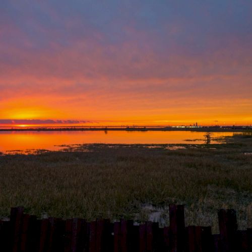 The image shows a sunset over a serene body of water, with grassy marshland in the foreground and a vibrant orange and purple sky in the background.